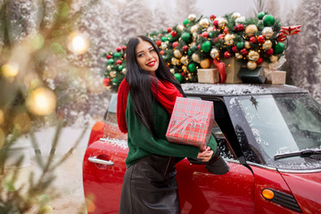 Pretty young girl is holding gift box near red car with decorated xmas tree on the roof, holiday concept.