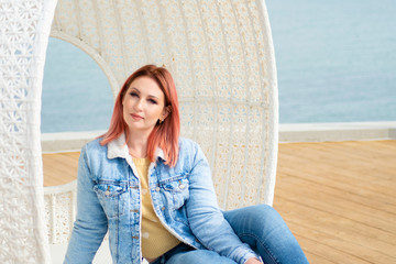 young woman with red hair and freckles is resting in swing, armchair near seashore