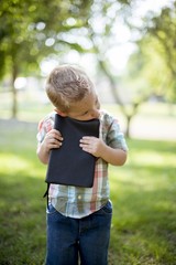 Vertical shot of a child holding the bible against his chest with a blurred natural background