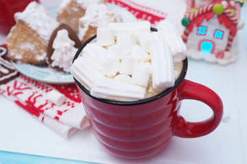 red mugs with coffee and marshmallows, gingerbread with icing in the shape of a snowflake on a red and white towel, red gift box, toys house and a snowman...