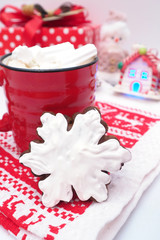 red mugs with coffee and marshmallows, gingerbread with icing in the shape of a snowflake on a red and white towel, red gift box, toys house and a snowman...
