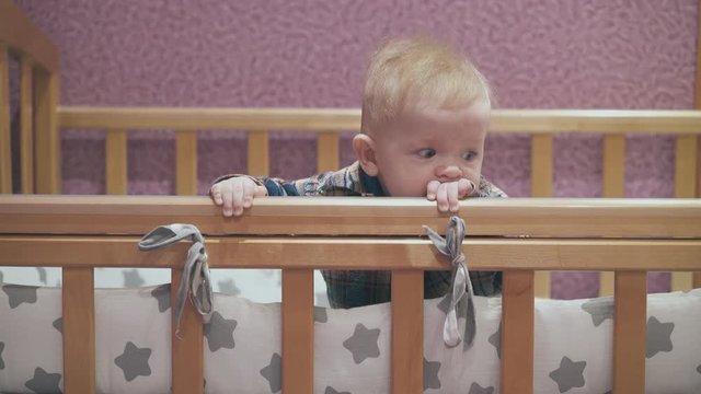 funny little boy leans on edge of wooden cot with star patterned linen against pink wall in light room closeup