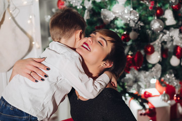 Son gives a Christmas gift to mom. Mom pleasantly surprised and cute looks at the child. Isolated on a New Year tree background with gifts, a fireplace and a homely atmosphere