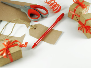Handmade gift tag carved from craft paper with red handle next to scissors, paper and ribbon offcuts and wrapped gift boxes. On light background with space for text. Handmade and celebration concept.