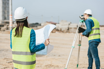 Selective focus of surveyor with blueprint and colleague using digital level on construction site - obrazy, fototapety, plakaty