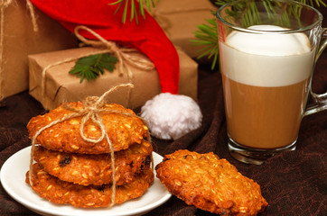 Christmas crispy oatmeal cookies, a transparent mug of coffee with milk or cappuccino, gift boxes in kraft paper, a hat forgotten by Santa Claus on a brown background. Holidays concept. Closeup