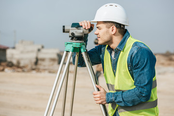 Side view of surveyor in hardhat looking throughout digital level on construction site