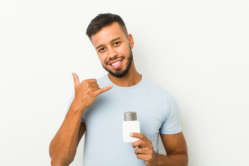 Young south-asian man holding an after shave cream showing a mobile phone call gesture with fingers.