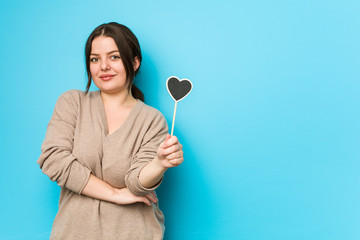 Young plus size curvy woman holding a heart shape smiling confident with crossed arms.