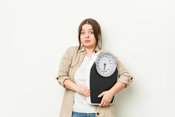 Young curvy woman holding a scale shrugs shoulders and open eyes confused.