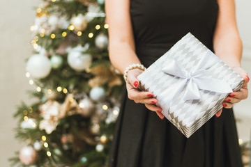 A girl in a festive black dress on the background of a Christmas tree with bokeh holds a gift and gives it to the camera. A woman gives a New Year's gift