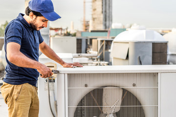 a professional electrician man is fixing a heavy duty unit of central air conditioning system by his tools on the roof top and wearing  uniform and white cap