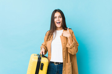 Young caucasian woman holding a suitcase surprised pointing at himself, smiling broadly.