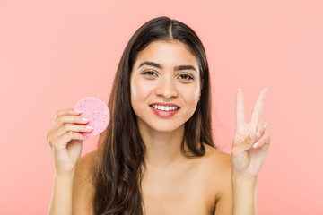 Young indian woman holding a facial skin care disc showing number two with fingers.