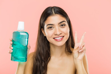 Obraz na płótnie Canvas Young indian woman holding a mouthwash bottle showing victory sign and smiling broadly.