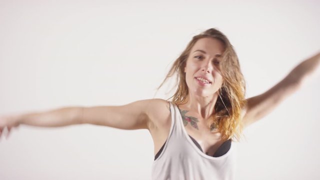 Waist up studio shot of young beautiful sweaty woman with wet hair and tattoos on chest looking at camera and performing seductive dance against white background
