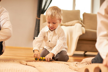 Obraz na płótnie Canvas Indoor shot of adorable Caucasian boy with blonde hair sitting on floor in living room with wooden railroad, holding toy train, having absorbed interested facial expression. Children and entertainment