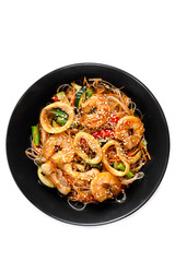 grilled king prawns, squid rings and vegetables mix, noodles in sauce, red pepper, sesame seeds in a black plate isolated on white background, top view