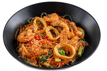 grilled king prawns, squid rings and vegetables mix, noodles in sauce, red pepper, sesame seeds in a black plate isolated on white background, side view