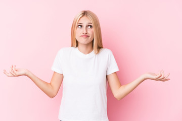 Young blonde woman on pink background doubting and shrugging shoulders in questioning gesture.