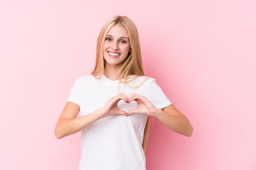 Young blonde woman on pink background smiling and showing a heart shape with hands.