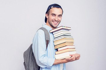 Happy student guy with backpack carries a stack of books on gray background, copy space, toned