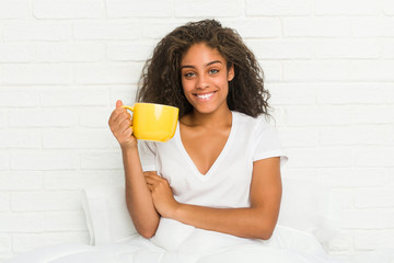 Young african american woman sitting on the bed holding a coffee mug smiling confident with crossed arms.