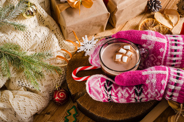 Hot Christmas and New Year's drink of cocoa with marshmallows. Hands in mittens are holding a mug with a drink on the table with gifts and Christmas tree branches.