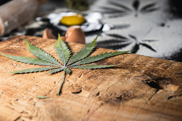 traces of marijuana leaves on flour, a broken raw egg, a rolling pin, and dried cannabis leaves on a wooden cutting board