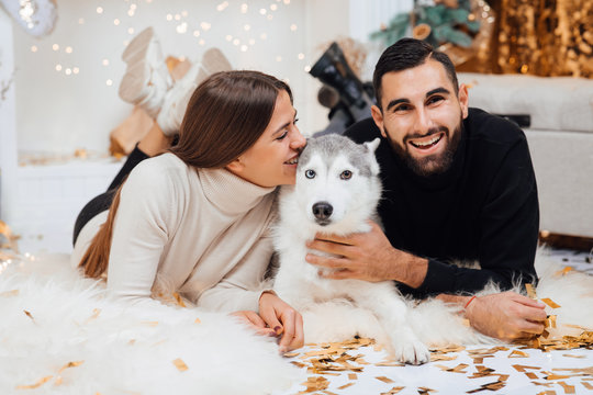 Merry Christmas and Happy New Year. Young beautiful couple are playing with their dog of the Husky in festive New Year living room just before the Christmas. Animal, people, holiday concept
