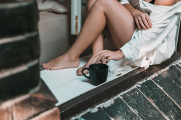 Woman in white dress is sitting on the windowsill with big black tea cup. Morning concept.