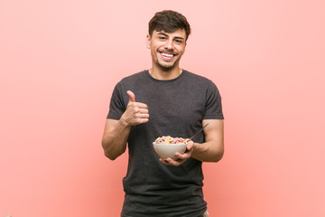 Young hispanic man holding a cereal bowl smiling and raising thumb up