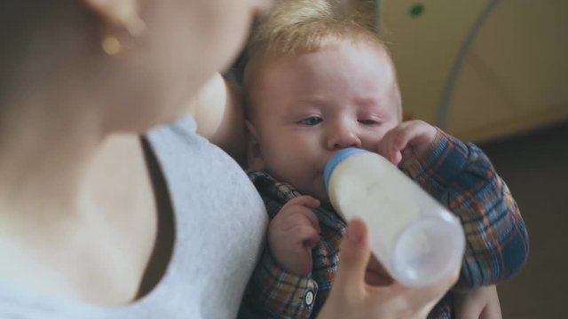 happy mommy rocks little son eating milk mix from plastic bottle with teat in arms against spacious light room close view