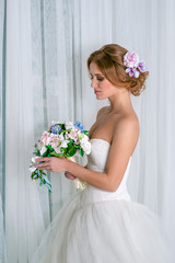 Studio shot of the calm bride  with bouquet of flowers wearing white dress and decoration in hair by the  window