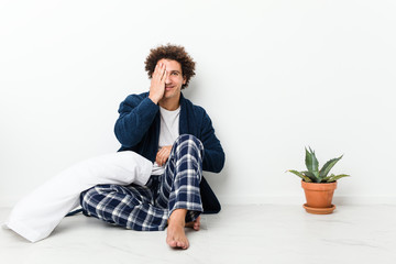 Mature man wearing pajama sitting on house floor having fun covering half of face with palm.