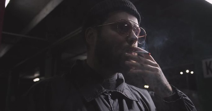 Young male hipster smoking cigarette in darkness outdoors, alone in street