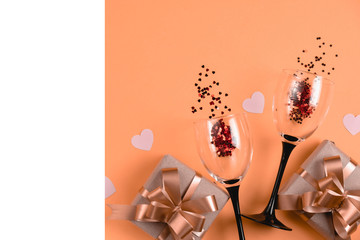 Holiday background. Two clinking wine glasses, handmade gift boxes decorated with pink heart confetti, white frame on orange background. Valentines day concept. Flat lay, top view, copy space