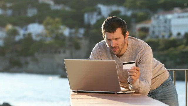 Worried man paying online with laptop and credit card in a balcony on the beach