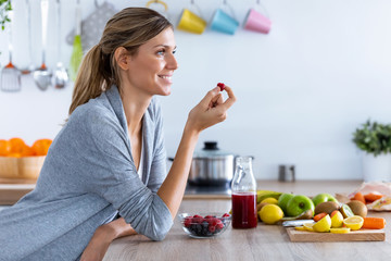 Pretty young woman eating red berries while sitting in the kitchen at home.