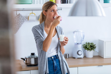 Pretty young woman eating yogurt while standing in the kitchen at home.