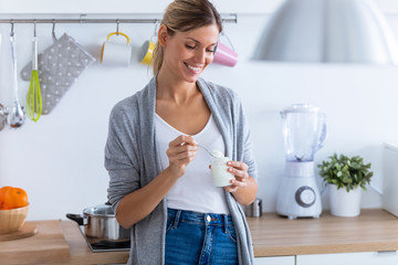 Pretty young woman eating yogurt while standing in the kitchen at home. - Powered by Adobe