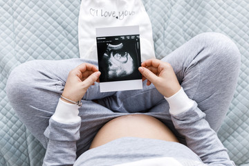 Expecting pregnant woman touching her belly while looking at ultrasound scans, sitting on cozy bed with blankets, pillows, kid clothes. Cropped image top view.