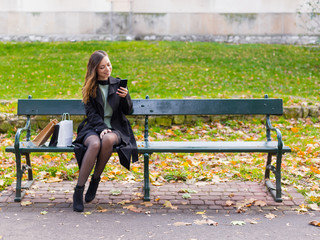 Beautiful girl sitting on a bench, woman with your phone writes a message on social networks, sunny autumn day in the park, woman relaxing after shopping.