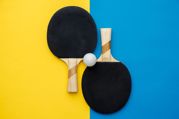 Two table tennis or ping pong rackets and ball on blue and yellow background. Top view. Copy, empty space for text