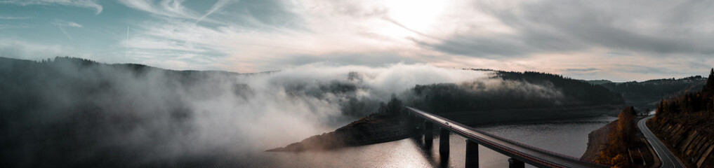 Aerial drone view from above of a morning mountain landscape with a lake dam and heavy low fog clouds hanging over the water and hills and winter mood.