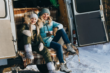 Two girls in winter clothes are sitting in the luggage compartment of the car during the day, holding a mug, looking at the camera and smiling against the background of a brown plaid.