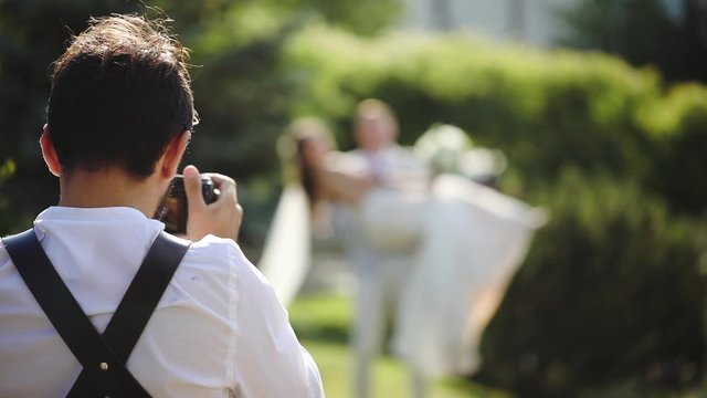 Back view of guy with camera shoots happy newlyweds on green city garden slow motion. Wedding photography, backstage