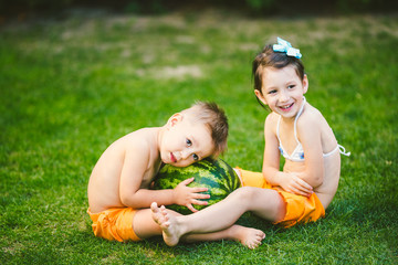 Two children, Caucasian brother and sister, sitting on green grass in backyard of house and hugging big tasty sweet watermelon berry, green striped color. Theme healthy food and ecological product