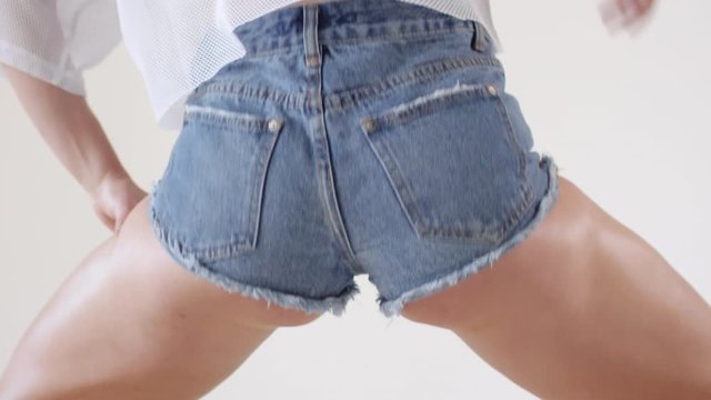 Rear view close up shot of sexy female dancer in jeans shorts shaking her booty while twerking against white background in studio