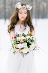 Beautiful bride. Winter wedding. Young woman in wedding dress with a floral cotton head wreath and bouquet standing in the winter forest and looking at the camera